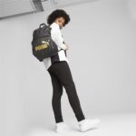 Person wearing a white top and black pants carries a black Puma backpack with a gold logo, standing against a gray studio backdrop.
