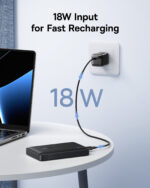 Laptop plugged into a small black power bank on a white round table, cable running to the wall outlet, text reads '18W Input for Fast Recharging'