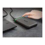 Dark gray external hard drive connected to a laptop via USB; a finger presses the power button with a green accent on the edge.