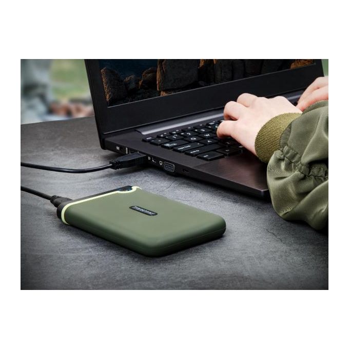 Green rugged external hard drive connected to a laptop via USB as someone types on the keyboard on a gray table.