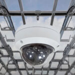 Dome surveillance camera mounted on a metal frame against a blue sky backdrop, surveillance setup visible above.