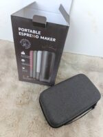 Box labeled 'Portable Espresso Maker' showing a compact espresso device illustration, placed on a beige rug beside a gray zippered carrying case.