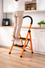 Person standing on an orange two-step ladder and reaching into an upper kitchen cabinet.
