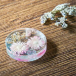 Round translucent resin ornament containing pink and white daisies, placed on a wooden table with a dried bouquet nearby.
